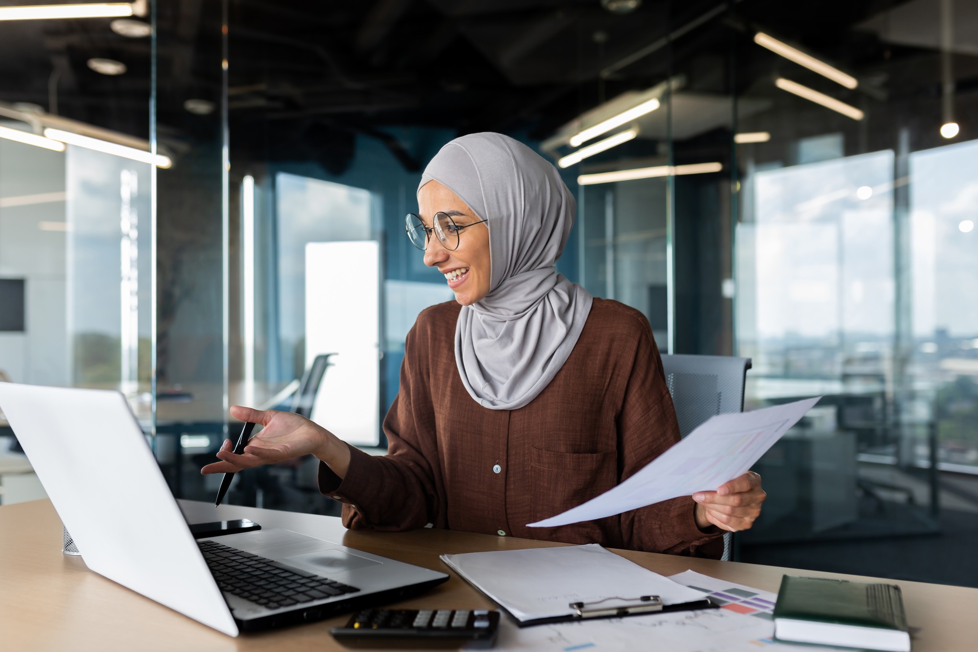 Young muslim businesswoman in hijab working in office with documents and talking on video call from